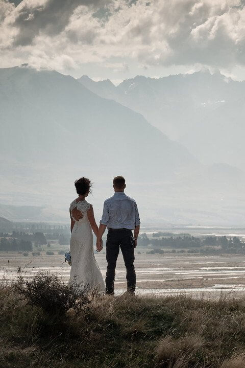 Rural wedding couple hold hands at White Rock, looking up Rangitata River to Two Thumb Range, New Zealand